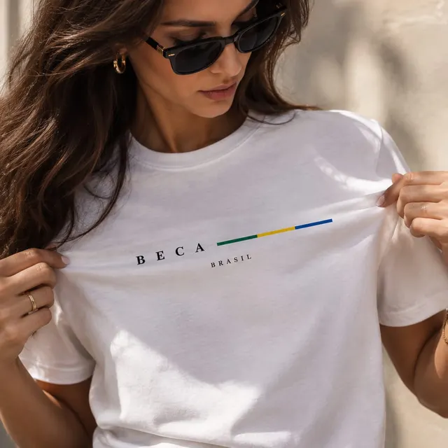 Mulher de óculos escuros vestindo camiseta branca com BECA e uma faixa verde, amarela e azul